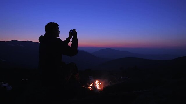 A man sits in the evening by a fire in the mountains and takes pictures of the night landscape on his smartphone. Tourist from Katra after sunset on the background of beautiful mountains.