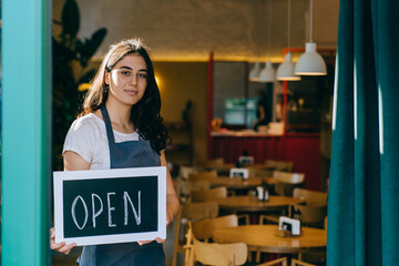 Young female owner or waitress in apron showing chalkboard with open sign at the coffee shop. Local business, hospitality, open after lockdown concept.
