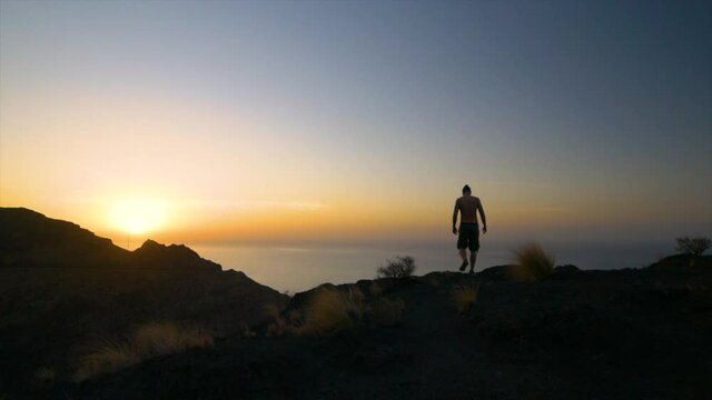 Silhouette of Man standing in Cliff next to ocean with Beautiful Sunset in Horizon then sits down to look at Sunset