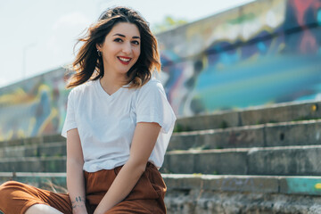 happy and tattooed woman looking at camera and sitting on stairs outside