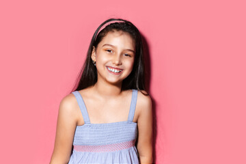 Studio portrait of happy little brunette child girl on background of pastel pink color. Wearing blue dress and headband.