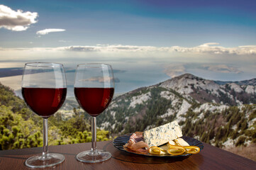 Two glasses of wine with charcuterie assortment on view of mountains and sea in Croatia. Glass of red wine with different snacks - plate with ham, sliced, blue cheese. Romantic celebration.