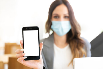 Female executive showing a smartphone with a blank screen at the office. She's wearing a face mask and formal wear. Selective focus. Space for text. Business and new normal concept. 