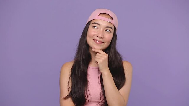 Young dreamful asian woman girl 20s in pink clothes cap posing isolated on pastel purple violet background studio. People lifestyle concept. Look around charming smile put hand prop up on chin think