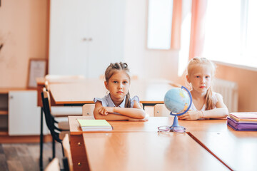 Obraz premium Two little schoolgirls sit at a desk in a school class and carefully listen to the teacher