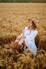 Stylish young girl in a summer white dress and hat posing in a golden wheat field. Fashion, glamour, lifestyle concept.