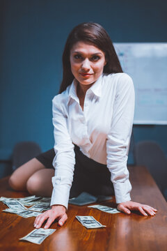 Beauty businesswoman sitting on office desk with paper money on it. Young girl in formalwear and scattered hundred dollar bills on table. Tinted image.