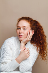 Portrait of a beautiful red-haired girl in a white knitted sweater on a beige background in the studio. The concept of natural beauty, no retouching, no makeup.