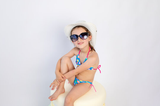 Cute little girl in black sunglasses and a blue bikini is sitting on the floor , turning sideways to the camera-Isolated on white background