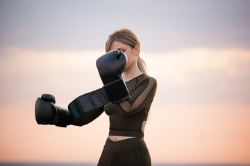 young adult athletic girl with long blond hair puts on black boxing gloves against the sunset sky