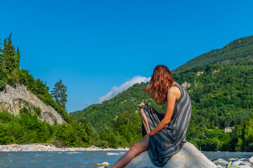 Fototapeta na wymiar An unrecognizable relaxed young Caucasian redhead woman sitting on a boulder with a beer bottle next to the Var river in the French Alps enjoying summer sunshine
