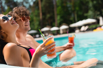 selective focus of cheerful girl in sunglasses holding cocktail near man in swimming pool