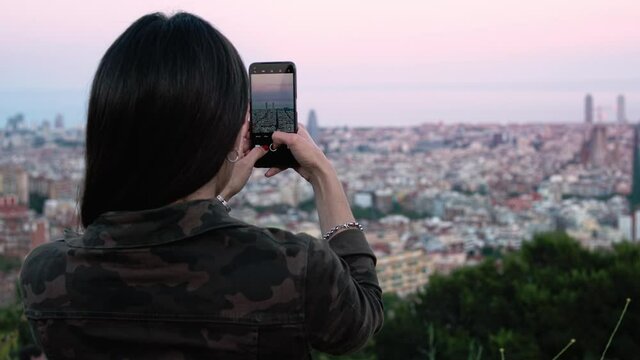 Young woman taking picture of Barcelona at sunset with smartphone device. Rear view of a girl standing at viewpoint enjoying cityscape at twilight. Mobile phone photography. Slow Motion
