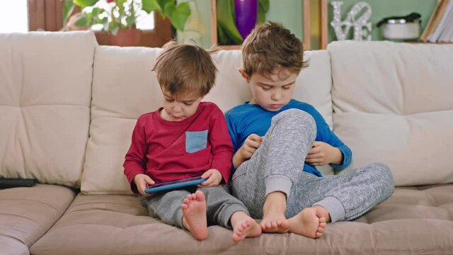 In front of the camera on the sofa two little kids brothers playing on the smartphone they spending the time together
