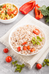 Unpolished rice porridge with stewed vegetables and oregano in white bowl on a gray concrete background. Side view, close up.