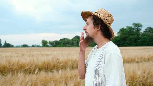 Stylish farmer of Caucasian appearance with a hat and beard walks in the field and looks at the wheat harvest