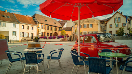 Terrace of street cafe with empty tables,wicker chairs and parasole,fenced off by red-and-white barrier tape,with red mini-car parked nearby,on German old square in center,Europe,on bright Sunny day