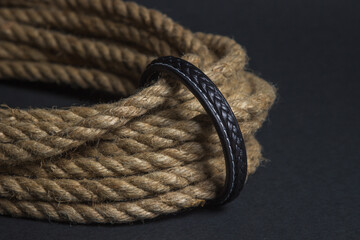 A man's leather bracelet is fastened to a rope. Male style on black background