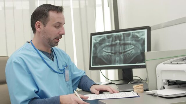 Lockdown of Caucasian male dentist wearing medical overall sitting at desktop at computer screen, explaining peculiarities of jaw x-ray while looking at camera