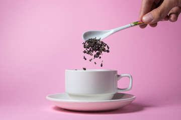 Tea leaves from a spoon are poured into a white porcelain cup on a pink background. Tea advertisement.
