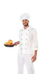 Handsome chef in uniform holding frying pan with ripe fruits and smiling at camera isolated on white
