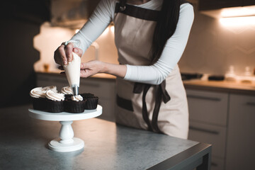Woman decorate chocolate cupcakes with whipped cream in pastry bag closeup in kitchen. Working at home.