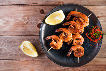 Appetizing fried shrimp on a skewer in a black plate on a wooden surface among lemon and seasonings. The concept of fast and wholesome food