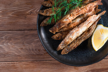 Appetizing fried capelin on a black plate stands on a wooden surface. Simple rustic food concept