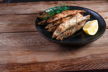 Appetizing fried capelin on a black plate stands on a wooden surface. Simple rustic food concept