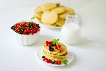 Summer breakfast pancakes with red and black currants in a bowl and milk in a glass jug on a white background.