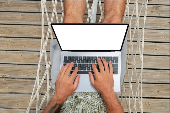 Fit young man lying in a net hammock working on laptop. Freelance remote work concept. Self employed man coding, wearing typical tourist shirt and hat. Copy space, sea view background - Powered by Adobe
