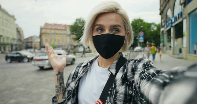 POV of Caucasian young beautiful stylish female hipster with blonde hair and in mask standing outdoors at street and talking via webcam. Close up of pretty joyful girl videochatting. Videochat concept