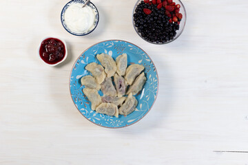 Boiled vareniki with blueberry and strawberry in blue plate on white wooden table, berries, jam, sour cream