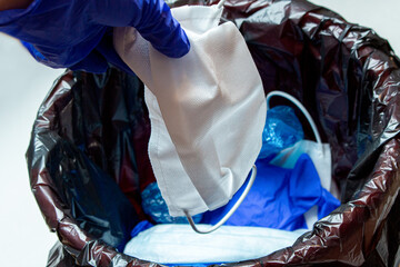 Hand in medical gloves throwing out discarded, used protective masks in the trash bin after quarantine