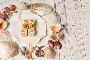 Fototapeta na wymiar Top view of Asian traditional Ebi Tempura Sushi in delivery takeaway box on sand background with Sea shells. Roll with black tiger shrimp, sesame seeds, Japanese mayonnaise and red caviar on top. 