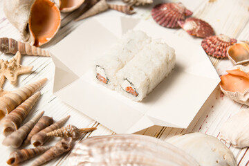 Japanese sushi roll with raw salmon and Philadelphia cream cheese wrapped in nori and rice in takeout delivery box. Sea shells and stars on white wooden board
