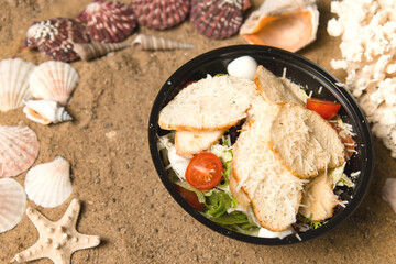 Caesar Salad with chicken fillet, cherry tomatoes, croutons, quail eggs and salad mix in black plastic bowl. Food Delivery package with meal. sea shells and stars on Sand background

