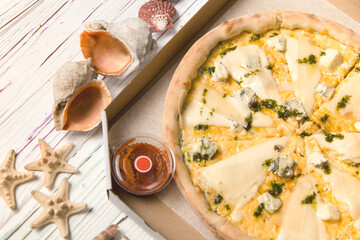 Four Cheese Uncut pizza in delivery box on parchment paper with and basil pesto sauce. wooden background with sea shells and stars. Pizza with Blue cheese, Gouda, Parmesan and Mozzarella. Tomato sauce