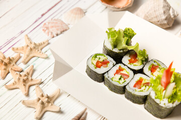 Japanese Futomaki Sushi Roll pieces on carton delivery box. Asian Vegetable Maki roll with tomato, cucumber, avocado, paprika, salad mix on wooden board with sea shells and stars on background
