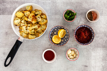 Pan with fried cauliflower florets and small bowls with other ingredients of Moroccan cooked salad Marak: homemade tomato passata, garlic, lemon, cumin. Top view, flat lay.