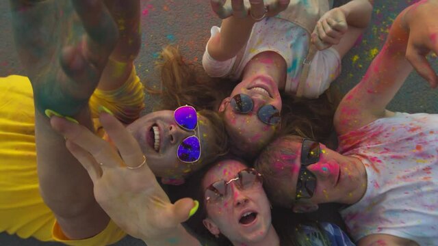 Happy friends in dirty clothes from colored powder and sunglasses, lying on the floor raising their hands to the top and smiling