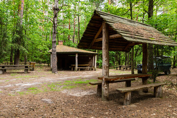 Fototapeta na wymiar Tourist recreation and rest place in forest. 4x4 offroad car parked next to picnic tables and wooden roof shelters behind trees. Kolacze, Poland, Europe.
