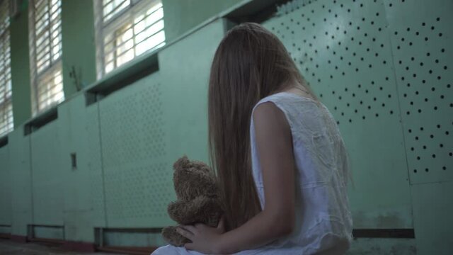 A girl is sitting in a white sundress in an old abandoned school gym. hair on his face, a toy in his hands. Horror.Abandoned house. Mysticism.