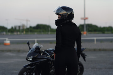 Biker woman in black leather jacket and full face helmet sits near stylish sports motorcycle at urban parking. Traveling and active lifestyle concept.