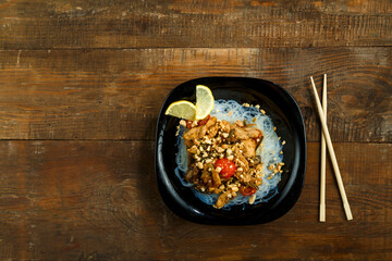 Nastol black plate with funchose and chicken with vegetables in pad Thai sauce next to chopsticks.