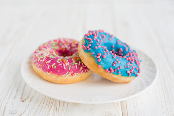 Two colorful assorted donuts on a small white plate with white background. Fresh sweet colorful homemade vanilla donuts on wooden background for birthday or party, top view, with free space for text