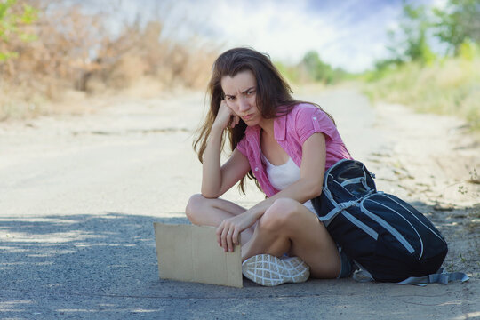 Hitchhiking. A Beautiful Young Woman With A Backpack And A Cardboard For Text Is Sitting On The Road And Is Sad Because She Cannot Catch A Passing Car To Get To The Place Of Travel.