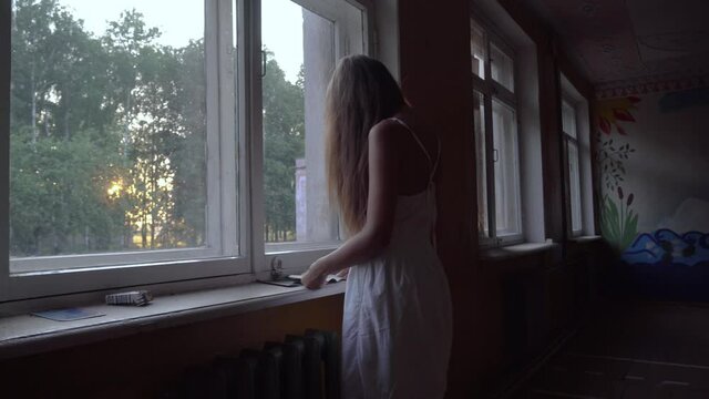 a girl in a white dress with long hair on her face stands near the window and examines objects on the windowsill. Interior of an abandoned house, school. post-apocalyptic ruins.