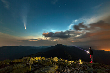 Silhouette of a hiker man with flashlight standing on mountain peak under starry sky lighting a...