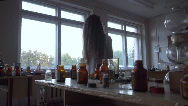 a girl with long black hair and a white dress walks slowly through the dark classroom of the school. The study of chemistry. Against the background of old test tubes and a blackboard.